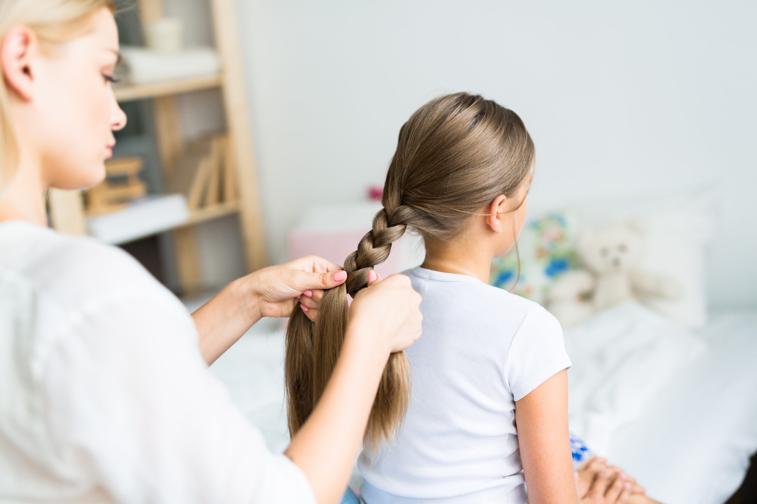 Mother French-braiding her daughter's long hair in a sunlit children's bedroom.