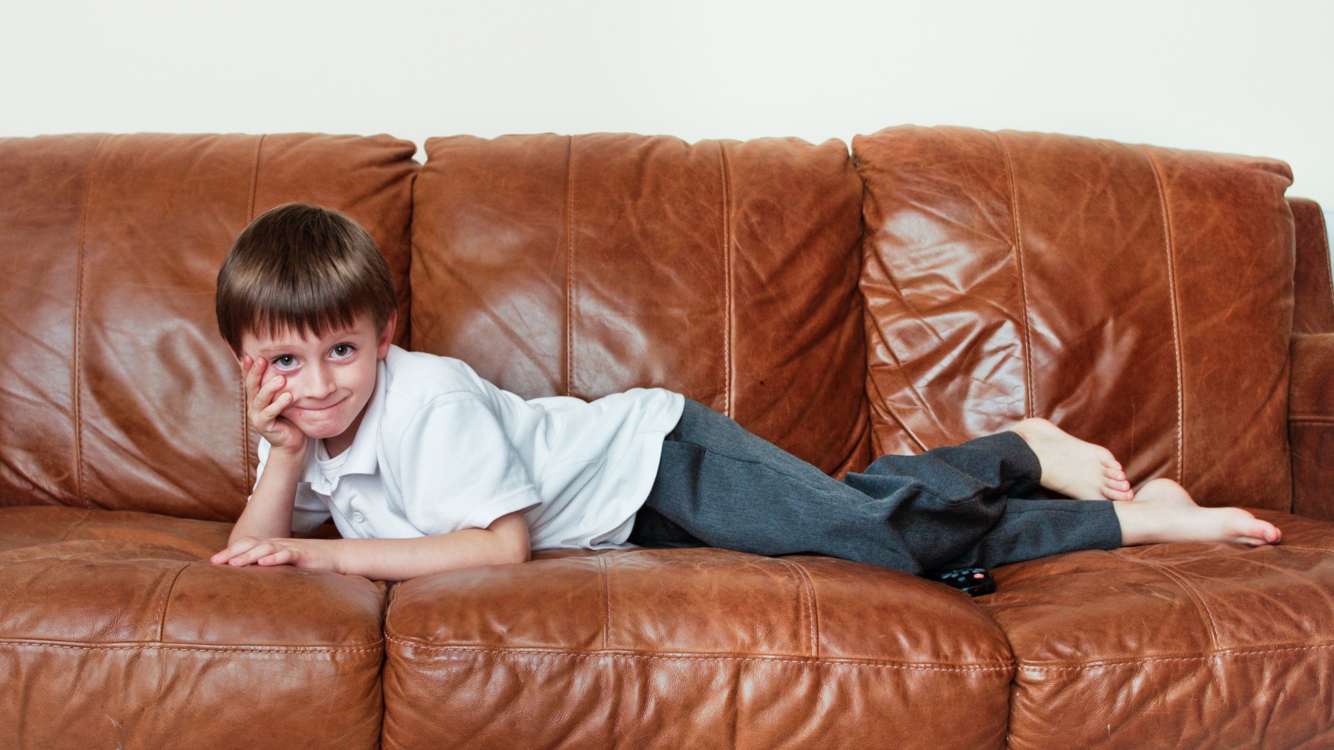 Calm mother in a sunlit Union County NJ living room wiping a brown leather sofa cushion with a microfiber cloth while her young daughter sits nearby, illustrating the parent decision to wipe leather surfaces rather than panic-clean after a head lice case