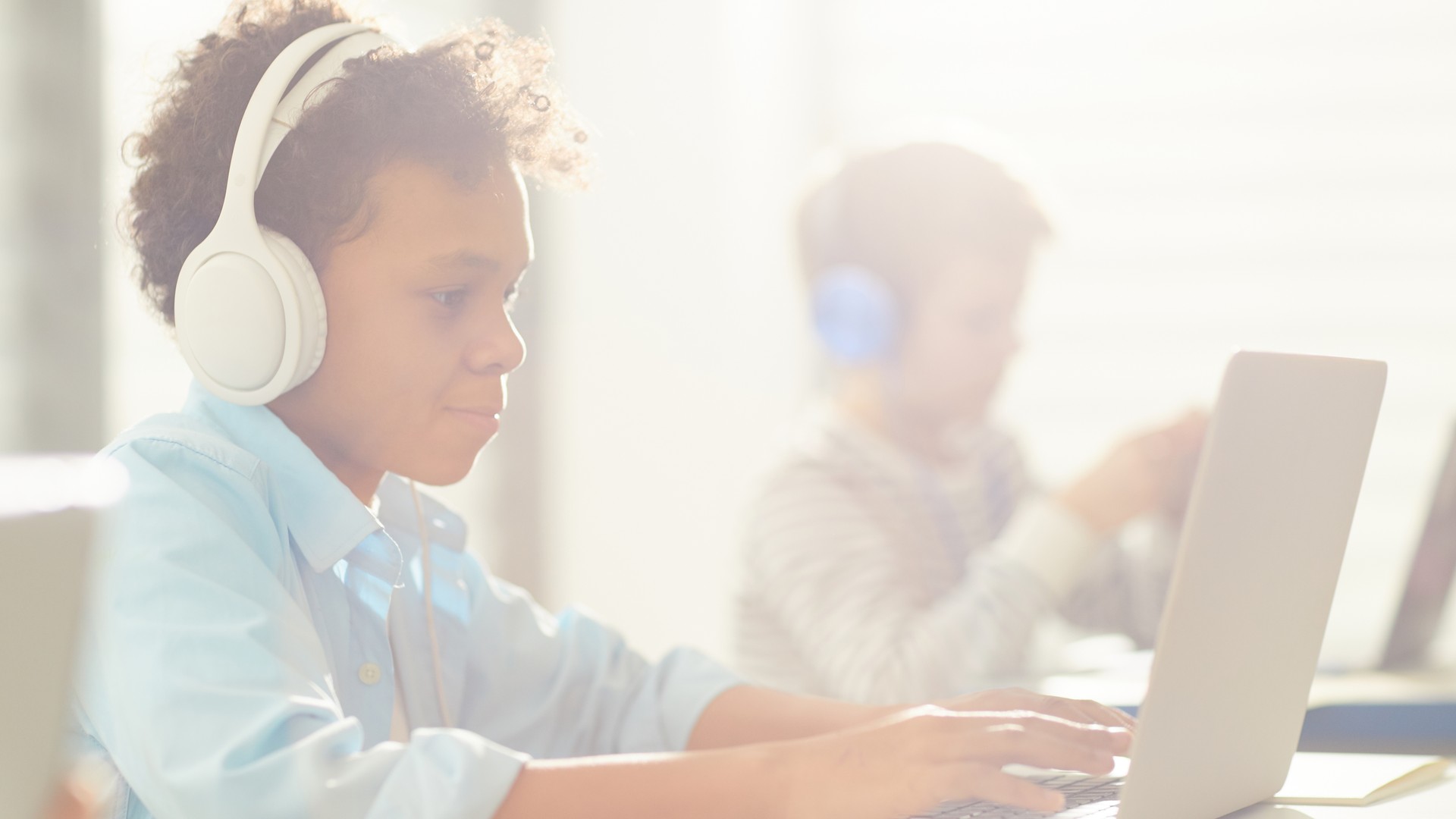 A grade-schooler at a classroom desk wearing over-ear headphones at a shared computer, next to a communal bin of headphones - the back-to-school moment behind the shared-headphones lice question