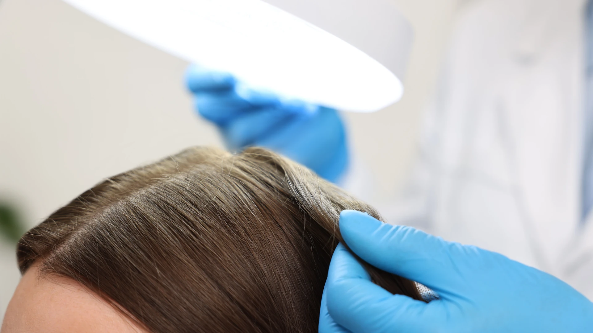 Blue-gloved hand separating brown hair beneath a bright light